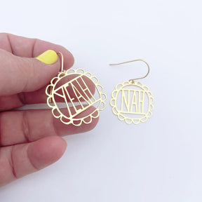 A pair of DENZ gold-toned oval dangle earrings with scalloped edges and cutout lettering "'YEAH NAH", shown on a white background. One earring is held by a hand with yellow nail polish, and the other hangs separately.