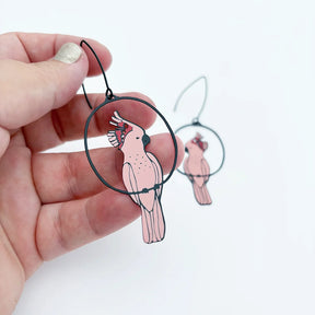 A hand holds a pair of DENZ Pink Cockatoo earrings with a second pair blurred in the background against a white backdrop.