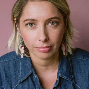 A close-up portrait of a woman with short blonde hair wearing DENZ Pink Cockatoo earrings and a denim shirt against a dusty pink background.