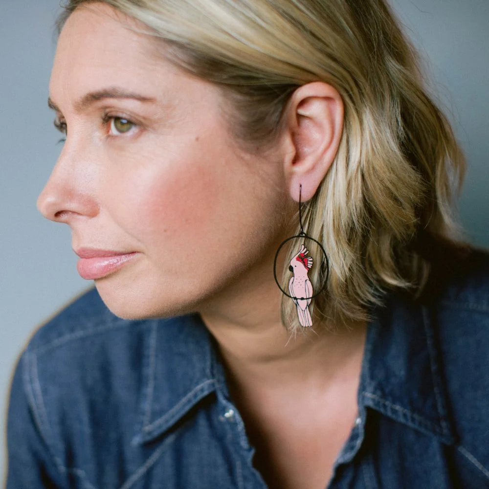 A close-up side profile of a woman with blonde hair wearing a DENZ Pink Cockatoo earring, paired with a blue denim shirt against a soft gray background.