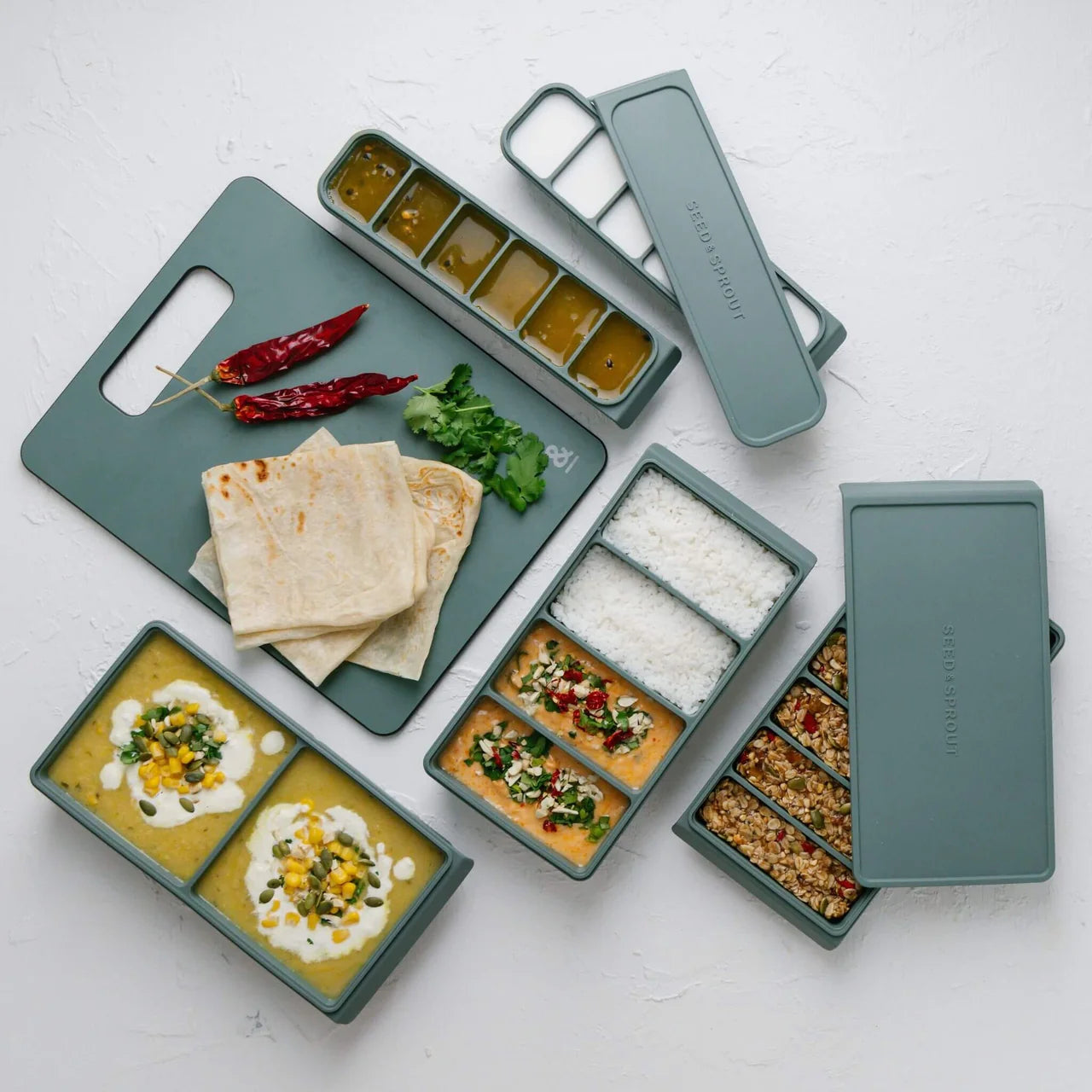 Seed & Sprout eucalyptus green silicone freezer trays with 4-portion and multi-compartment dividers, displayed alongside meal prep containers and a matching cutting board.