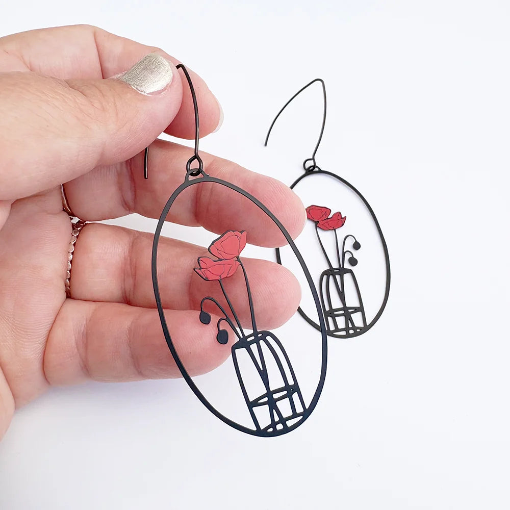A hand holds a pair of DENZ Poppy Vase earrings with red flower and green stem cutout details, shown against a plain white background.