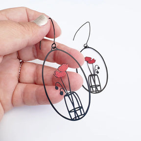 A hand holds a pair of DENZ Poppy Vase earrings with red flower and green stem cutout details, shown against a plain white background.