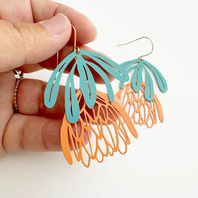 Hand holding a pair of DENZ Protea Flower dangle earrings in teal and orange with hand-drawn botanical design on a white background