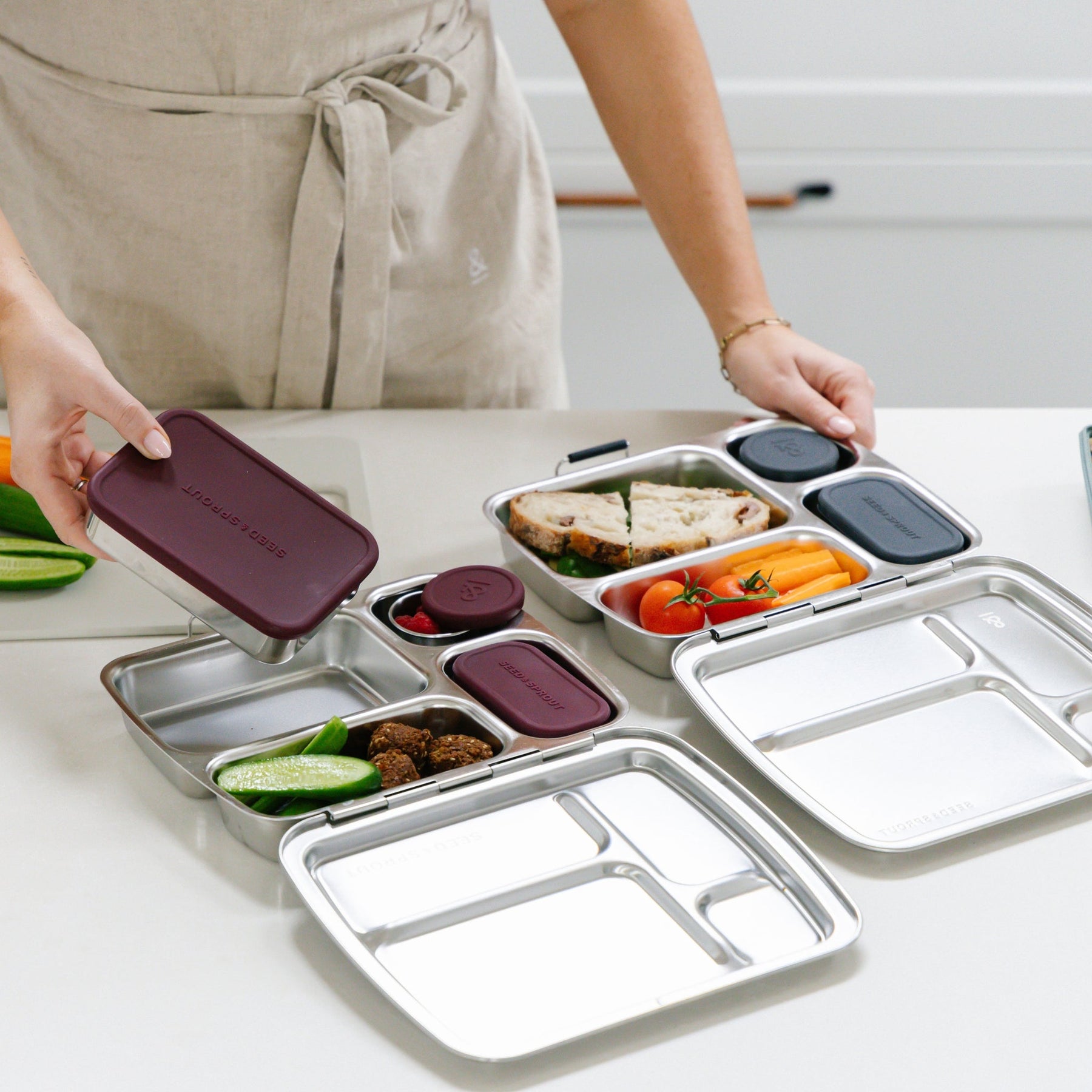 Seed & Sprout person packing a mulberry stainless steel CrunchBox bento lunch box with a small container, alongside another bento box filled with a sandwich and vegetables on a kitchen counter.