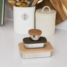Seed & Sprout stainless steel containers and leakproof snack pots in pecan tones, stacked on a white kitchen counter.
