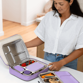 Woman opening a purple Seed & Sprout insulated lunch bag containing a stainless steel CrunchBox bento lunch box filled with fresh berries, with another bento box on the counter beside her.