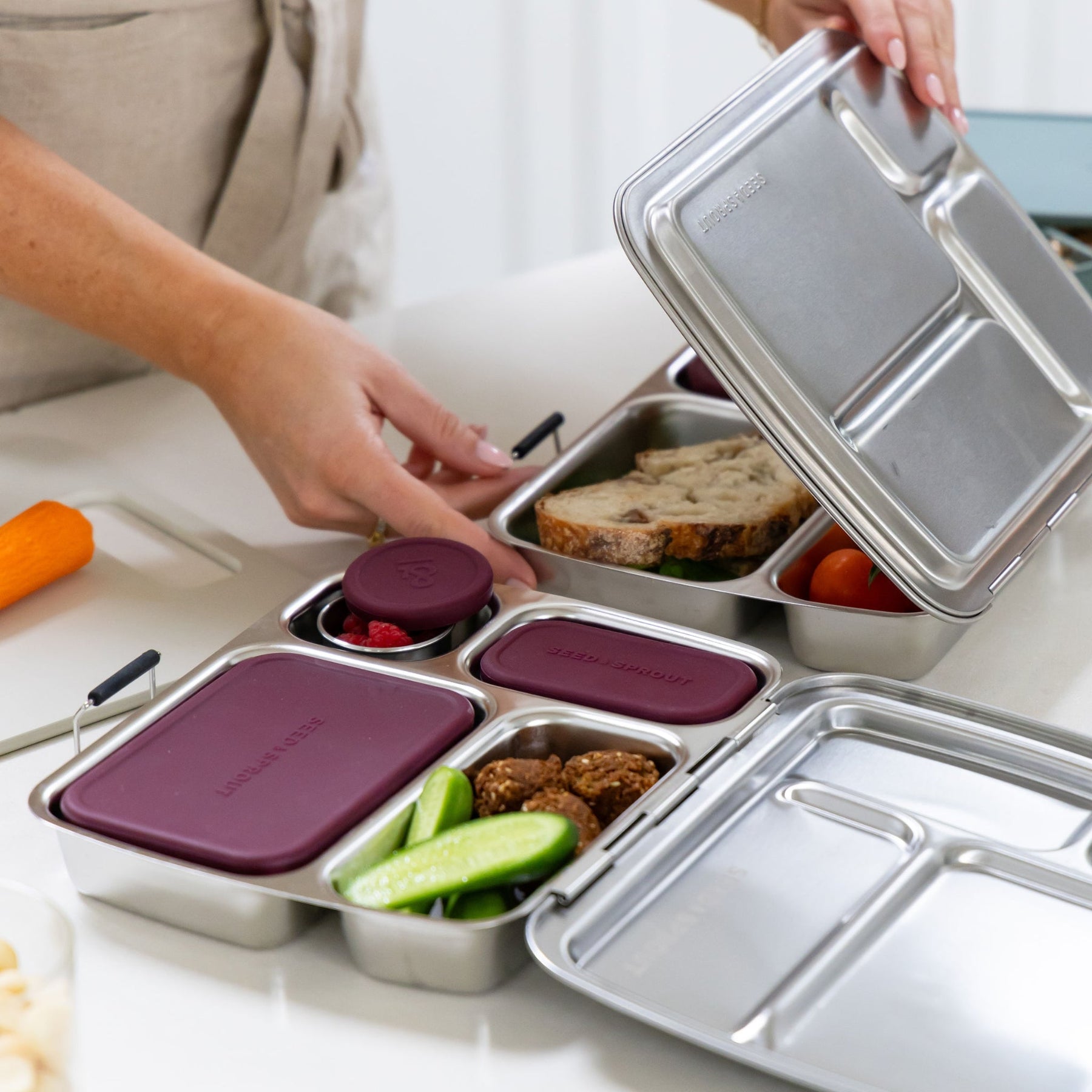 Seed & Sprout stainless steel CrunchBox bento lunch box in mulberry, packed with falafel, cucumber, and berries, with another lunch box containing a sandwich on the side.