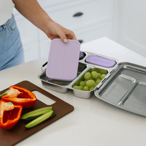 Person placing a plum silicone-lidded leakproof container into a Seed & Sprout stainless steel CrunchBox bento lunch box filled with green grapes, with sliced red capsicum and cucumber on a cutting board.