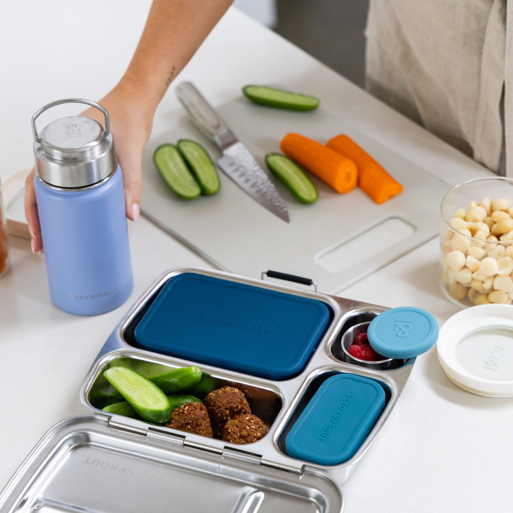 Seed & Sprout sea glass colour insulated water bottle alongside a Forest green CrunchBox bento lunch box filled with falafel, cucumber slices, and berries, set on a kitchen counter next to a cutting board with vegetables.