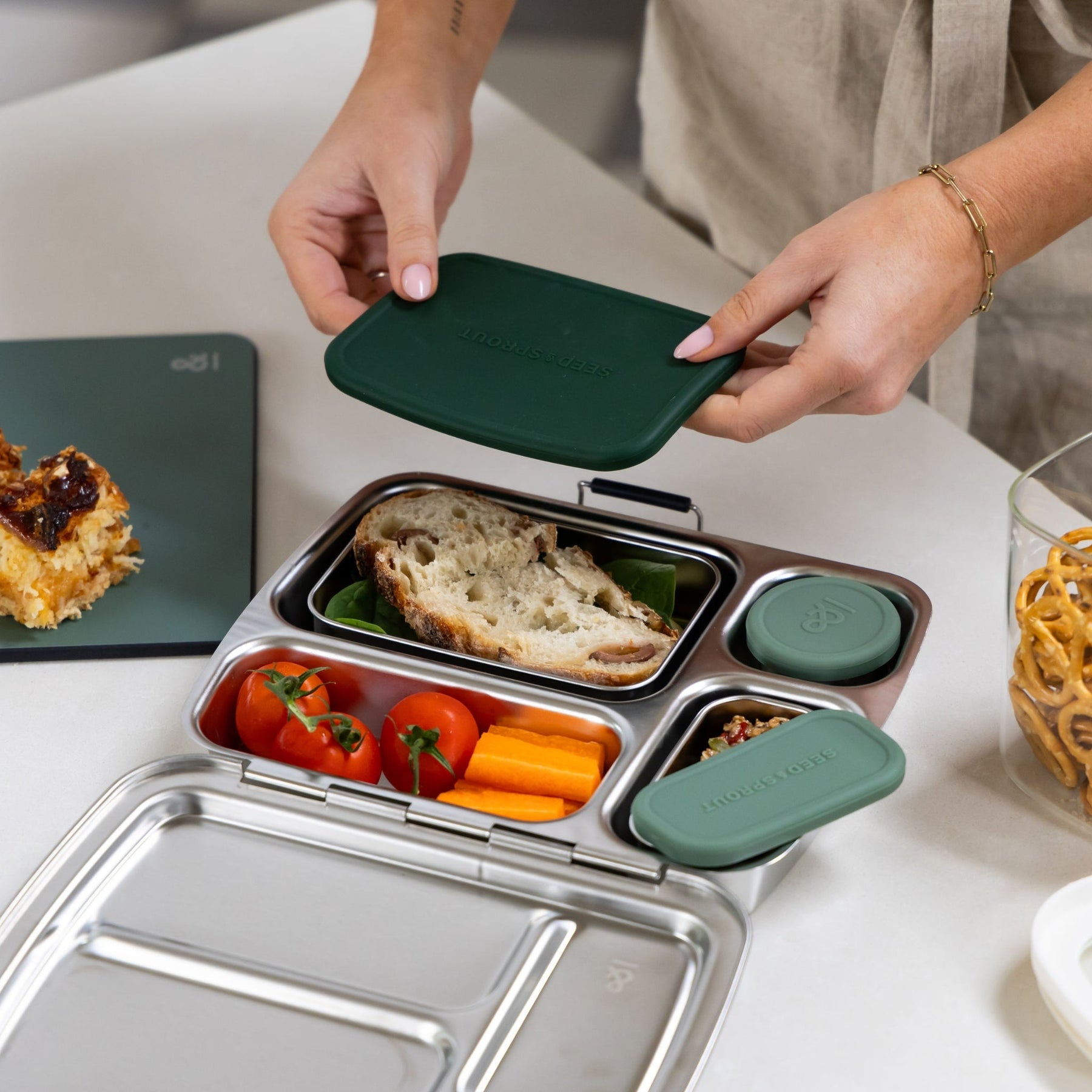 Seed & Sprout person packing a moss stainless steel CrunchBox bento lunch box with a sandwich lid, alongside fresh tomatoes, carrots, and snacks on a kitchen counter.