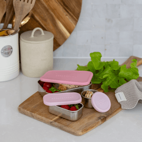 Seed & Sprout stainless steel containers with pink silicone lids and matching leakproof snack pots filled with fresh berries, arranged on a wooden cutting board in a kitchen setting.