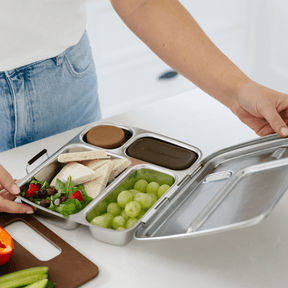Seed & Sprout stainless steel bento lunch box with partitioned compartments, a durable stainless steel design, and removable leakproof silicone pots, being opened by a person.