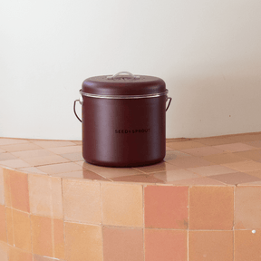 Seed & Sprout kitchen compost bin in rich mulberry color displayed centrally on warm terracotta-toned tiled surface.