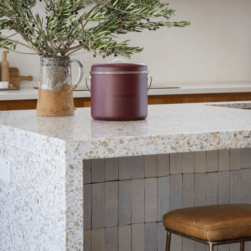 Seed & Sprout mulberry kitchen compost bin displayed on a modern terrazzo countertop next to an earthy ceramic vase filled with leafy branches.