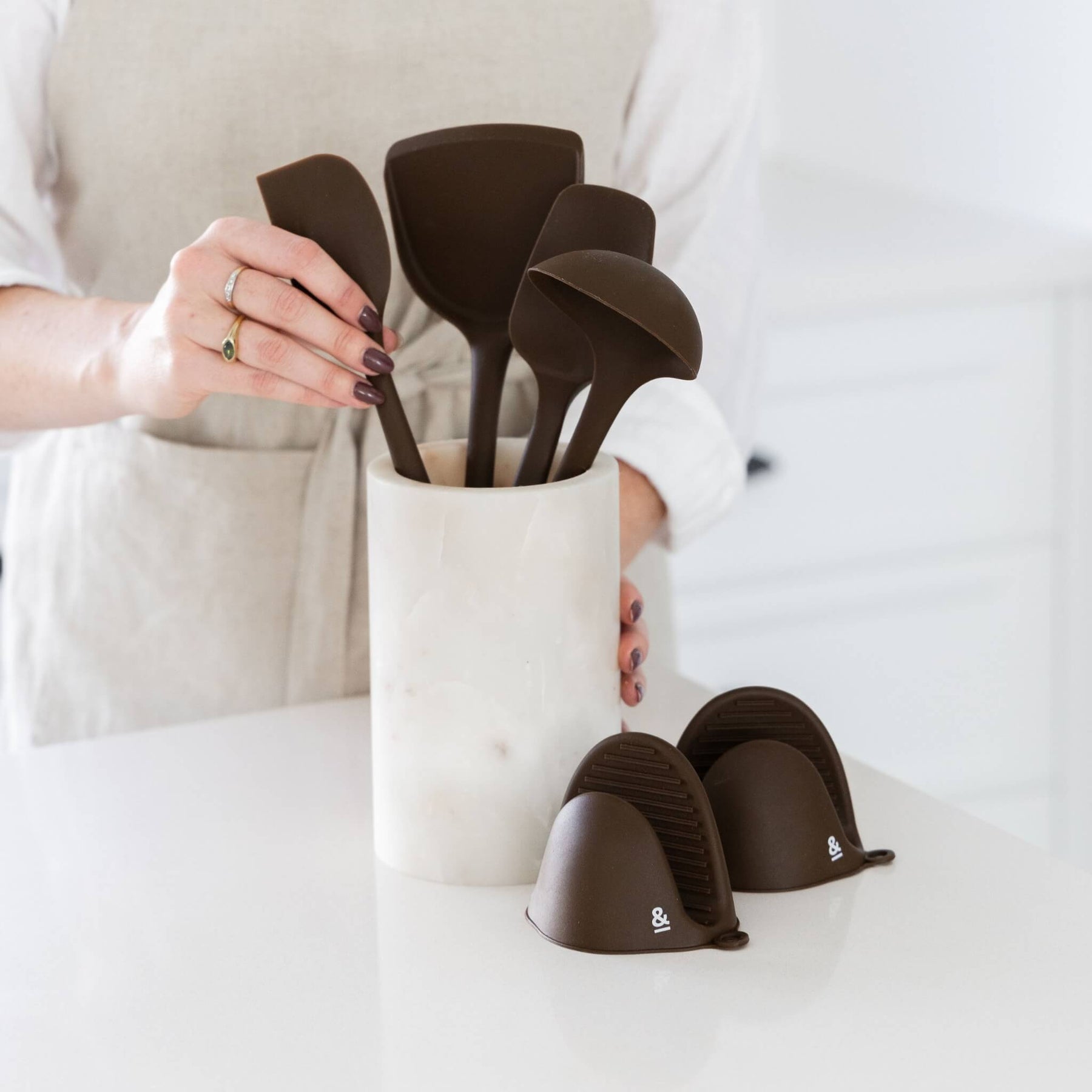 A person arranging Clove-brown Seed & Sprout silicone kitchen utensils in a marble crock next to matching mini pinch mitts on a kitchen counter.