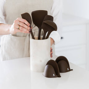 A person arranging Clove-brown Seed & Sprout silicone kitchen utensils in a marble crock next to matching mini pinch mitts on a kitchen counter.