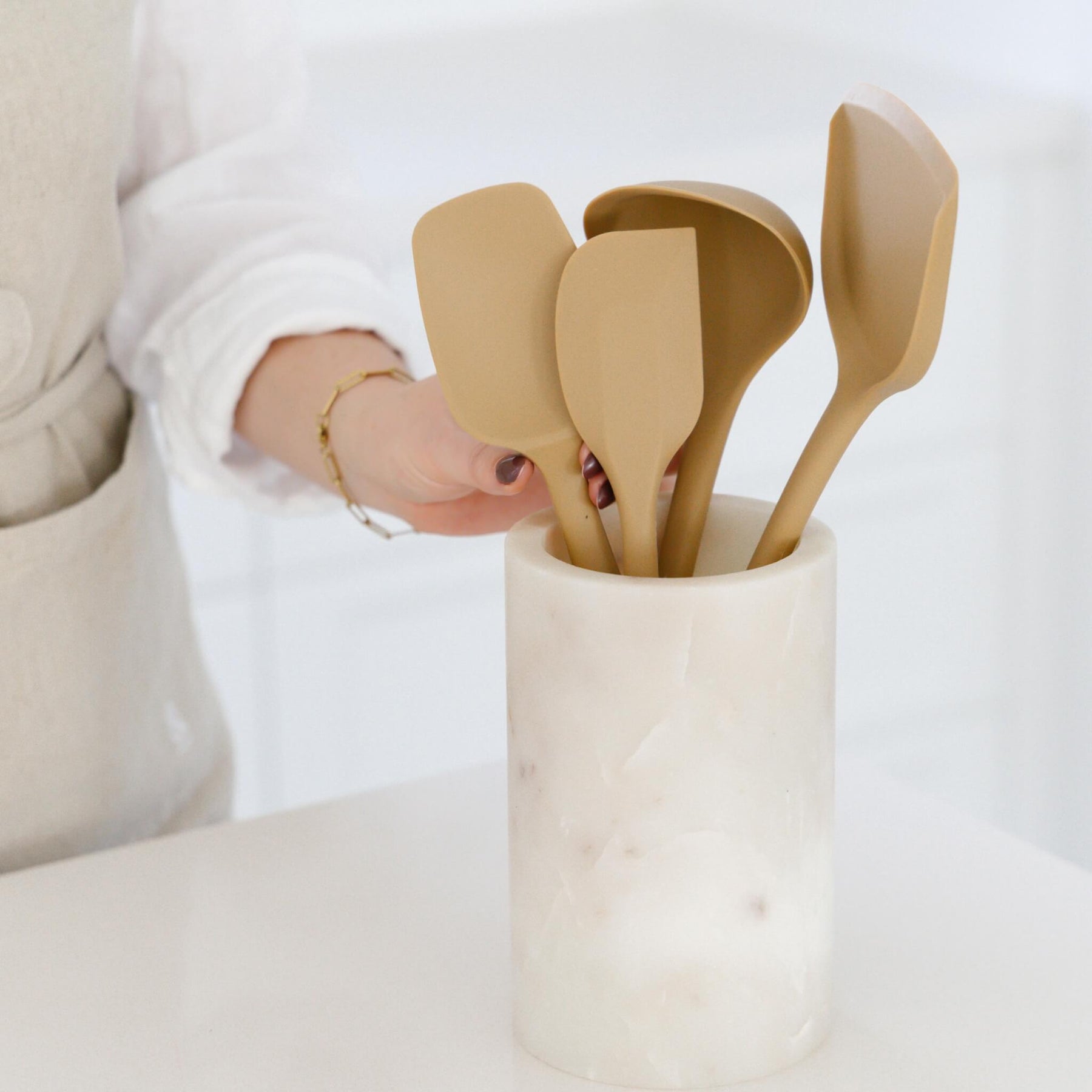 A person arranging a set of Praline-colored Seed & Sprout silicone kitchen utensils inside a marble crock on a kitchen counter.