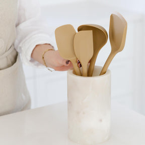 A person arranging a set of Praline-colored Seed & Sprout silicone kitchen utensils inside a marble crock on a kitchen counter.