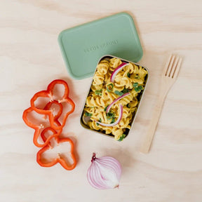 Seed & Sprout stainless steel CrunchPot container filled with pasta salad, displayed alongside its sage green silicone lid, red bell pepper slices, half a red onion, and a wooden fork.