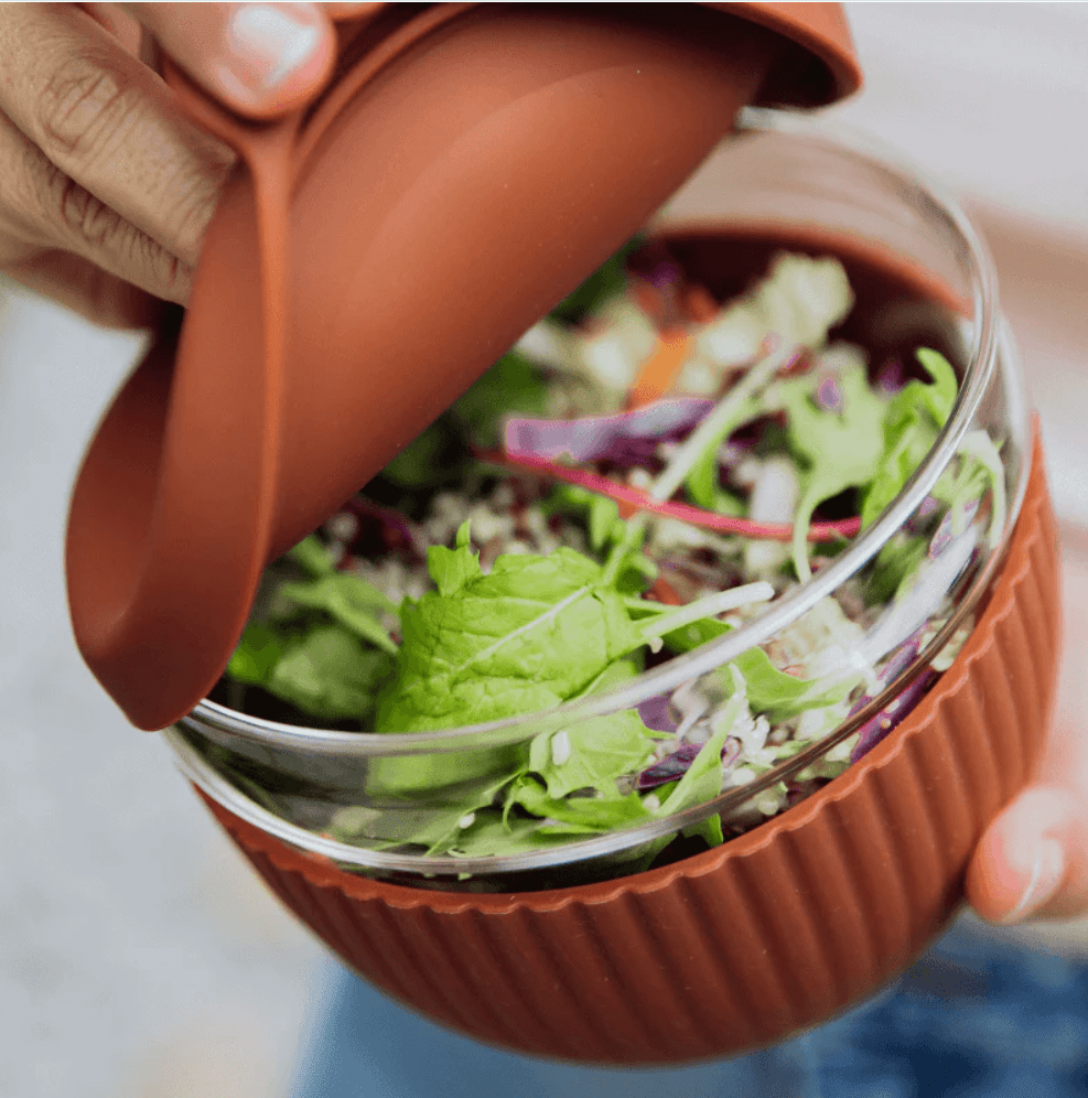 Seed & Sprout person opening a Cocoa  1000ml glass lunch bowl filled with a fresh green salad, highlighting its protective ribbed silicone base and matching leakproof lid.