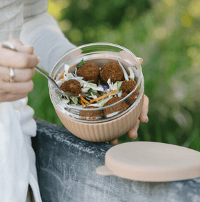 A person using a fork to eat a falafel and shredded vegetable salad from a Praline-toned 1000ml glass lunch bowl outdoors, with its matching leakproof silicone lid resting on a nearby surface.