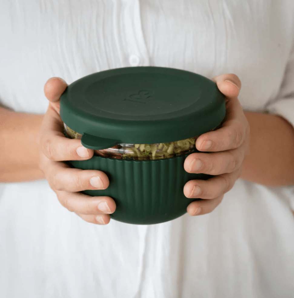 A person holding a sealed Seed & Sprout in Moss 1000ml glass lunch bowl filled with a fresh meal, highlighting the leakproof silicone lid and protective ribbed sleeve.