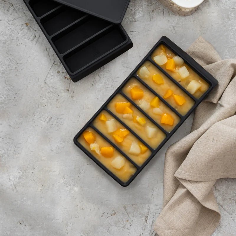Seed & Sprout charcoal silicone freezer-to-oven tray filled with fruit purée and chunks, with a matching lid and tray displayed on a textured stone surface.