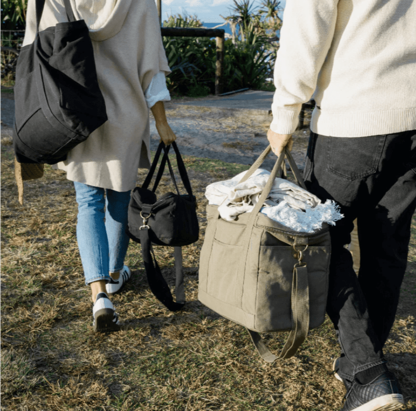 Two people walking outdoors carrying Seed & Sprout insulated cooler bags in olive and charcoal on a sunny day.