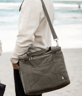 Seed & Sprout insulated cooler bag worn over the shoulder by a person walking along a sandy beach near the ocean.