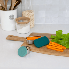 Seed & Sprout leak-proof stainless steel 2 pot set with eucalyptus green silicone lids, filled with carrot sticks and dip on a wooden cutting board.