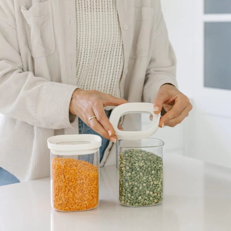 A person opening a Seed & Sprout Newrybar Square Glass Pantry Jars filled with green split peas next to another jar filled with red lentils.