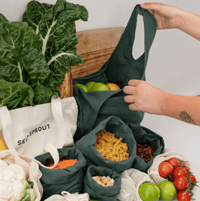 Hands holding a charcoal grey organic cotton shopping tote surrounded by Seed & Sprout bulk bags and mesh produce pouches filled with groceries.