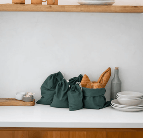 Set of charcoal green Seed & Sprout organic cotton bulk and bread drawstring bags displayed on a kitchen benchtop next to ceramic tableware.