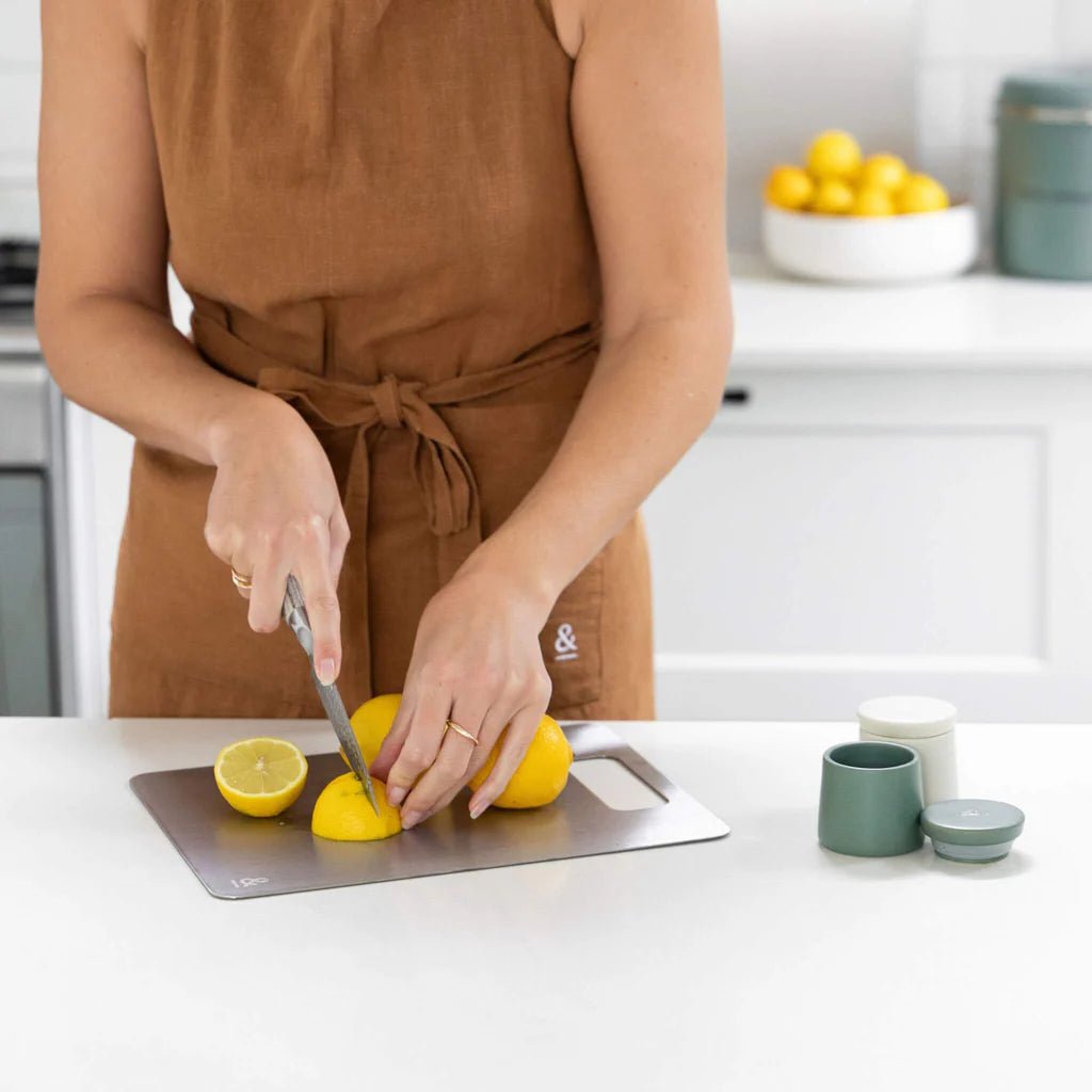 A person wearing a brown apron slicing lemons on a Seed & Sprout Titanium  steel cutting board on a white countertop.