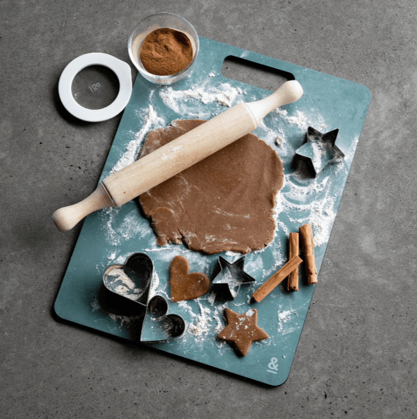Seed & Sprout Eucalyptus wood fibre chopping board set up for baking, featuring rolled cookie dough, a wooden rolling pin, star and heart-shaped cookie cutters, scattered flour, cinnamon sticks, and a small bowl of spice.