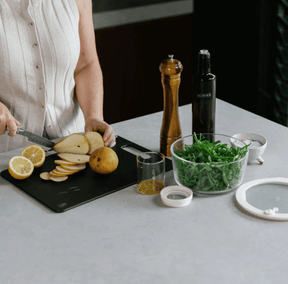 Seed & Sprout Liquorice wood fibre chopping board set up for food prep with sliced pears and lemons, accompanied by a wooden pepper grinder, olive oil bottle, and fresh salad greens on a kitchen counter.