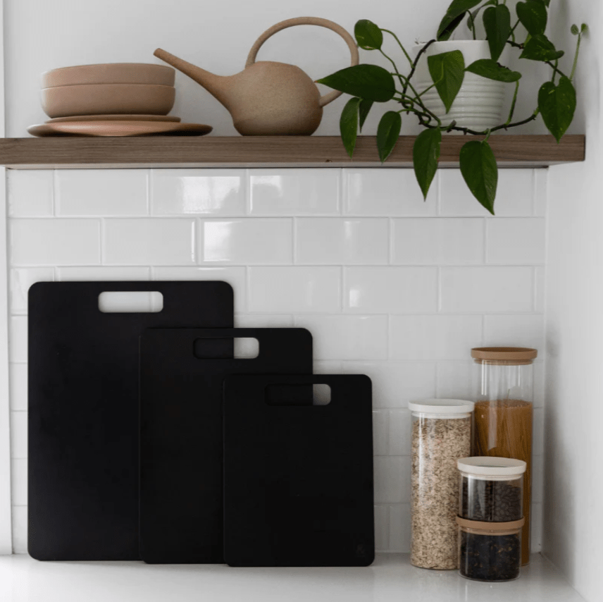 Seed & Sprout set of three Liquorice wood fibre chopping boards displayed in graduated sizes against a white tiled kitchen backsplash, alongside pantry jars and a styled shelf.