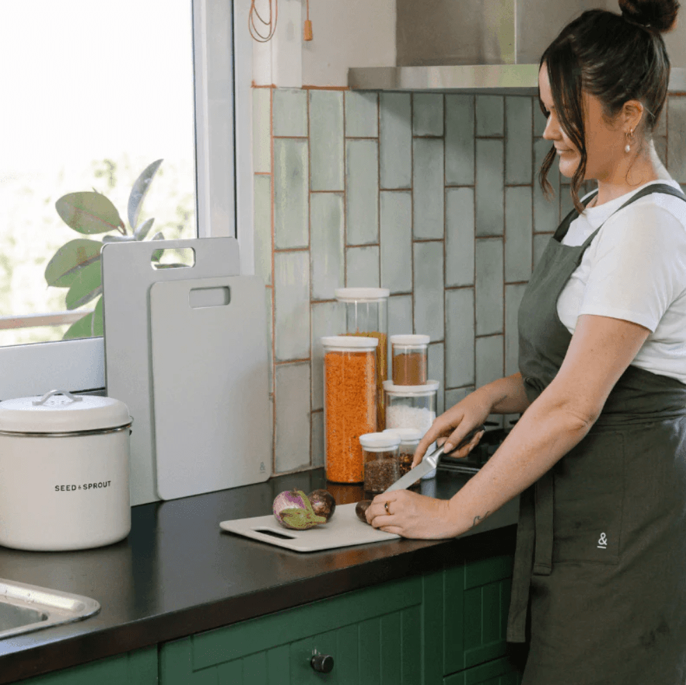 A person wearing a dark green apron chopping vegetables on a Seed & Sprout mushroom-colored wood fibre cutting board in a kitchen.