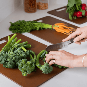 Seed & Sprout person chopping fresh broccolini on a Nutmeg wood fibre chopping board in a kitchen, with additional matching boards staged with whole carrots and radishes.
