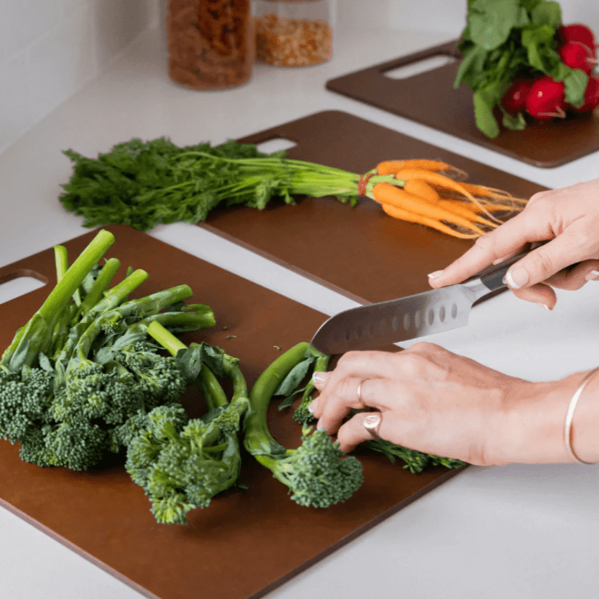 Seed & Sprout person chopping fresh broccolini on a Nutmeg wood fibre chopping board in a kitchen, with additional matching boards staged with whole carrots and radishes.