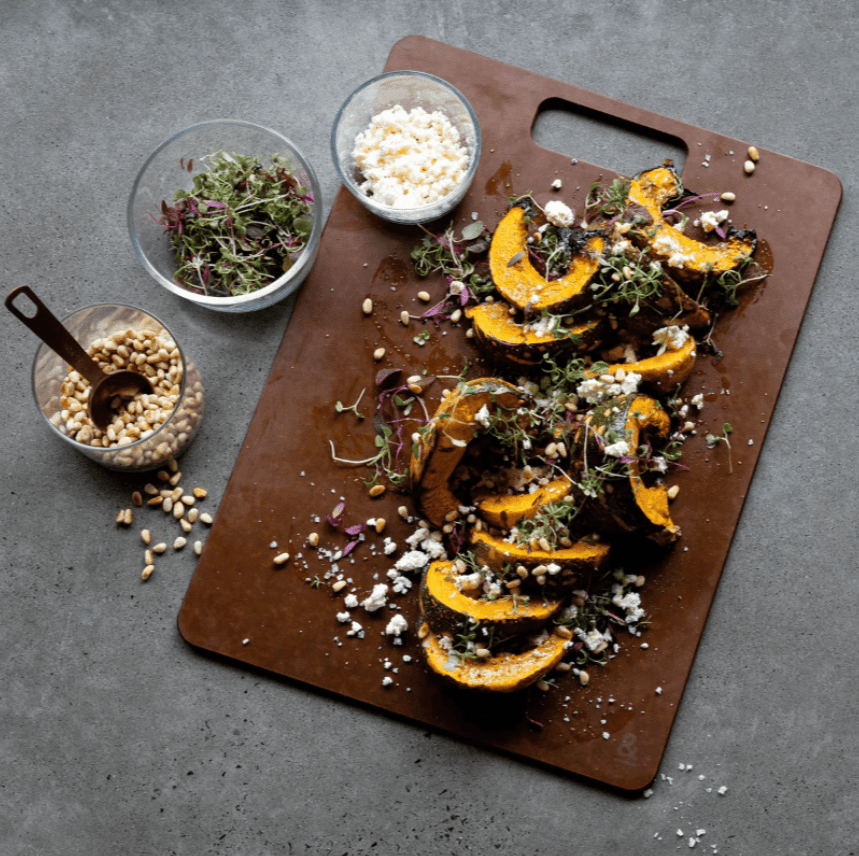 Seed & Sprout Nutmeg wood fibre chopping board arranged as a serving platter with roasted pumpkin wedges garnished with feta, microgreens, and pine nuts, alongside bowls of toppings on a dark countertop.