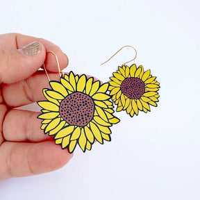 A hand holds a pair of DENZ Sunflower Earrings in Brown & Yellow against a plain white background. The earrings feature bright yellow petals, black outlines, and textured brown centers.