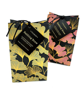Two Thurlby Herb Farm Gingko Bathing Brew packages standing side by side; one features a black paper bag with yellow-green ginkgo leaves, and the other features pink and gold ginkgo leaves, both tied with a black cord and branded tags.