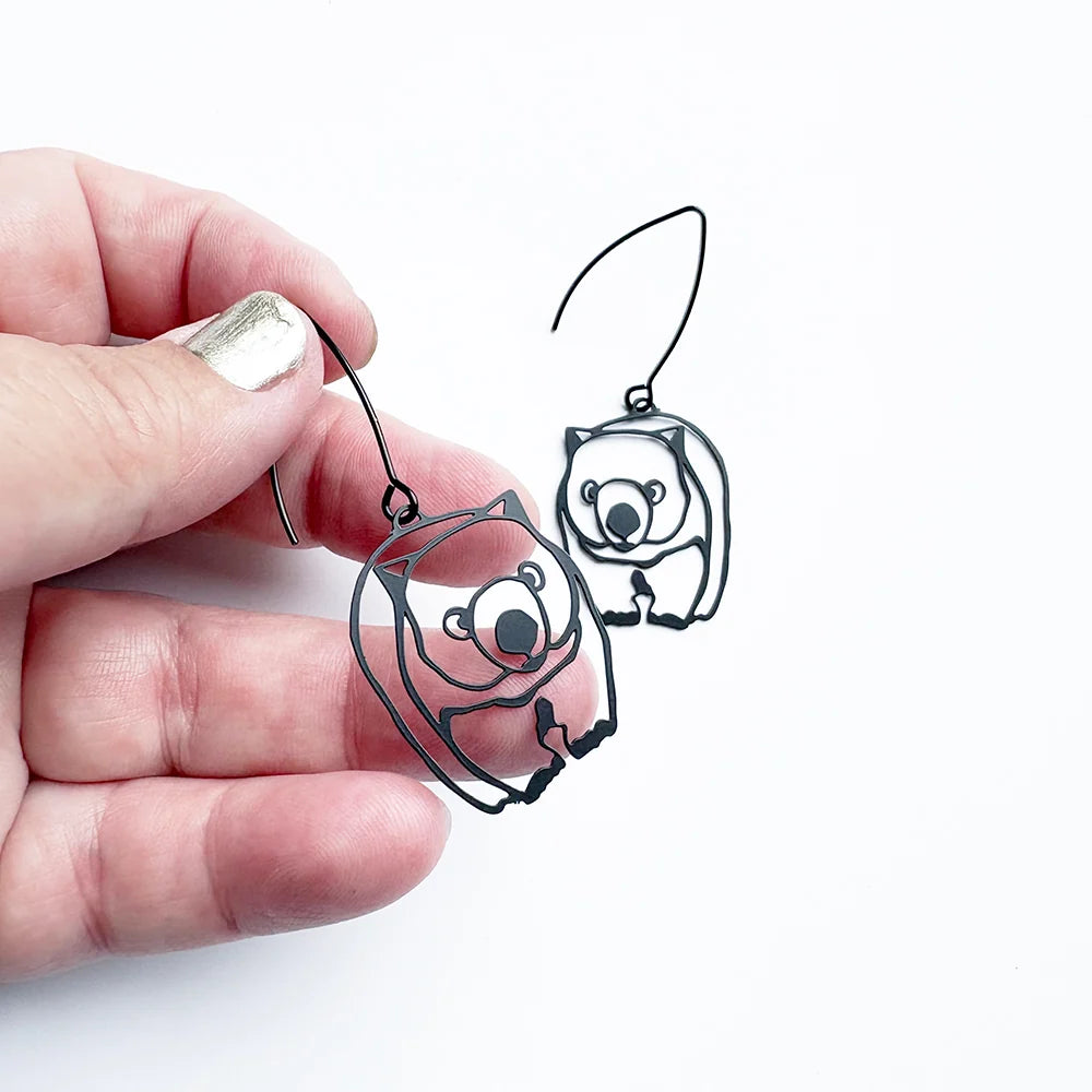 A hand holds a pair of DENZ Wombat  dangle earrings against a plain white background. One earring is shown hanging, and the other is held by the hand to display the design and hook.