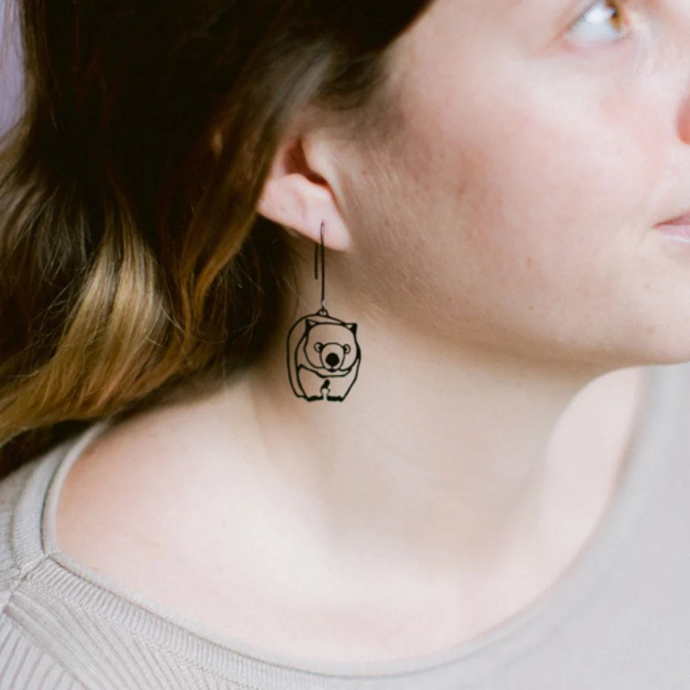 A close-up lifestyle shot of a woman wearing a minimalist DENZ Wombat dangle earring shaped like a small bear inside a square.