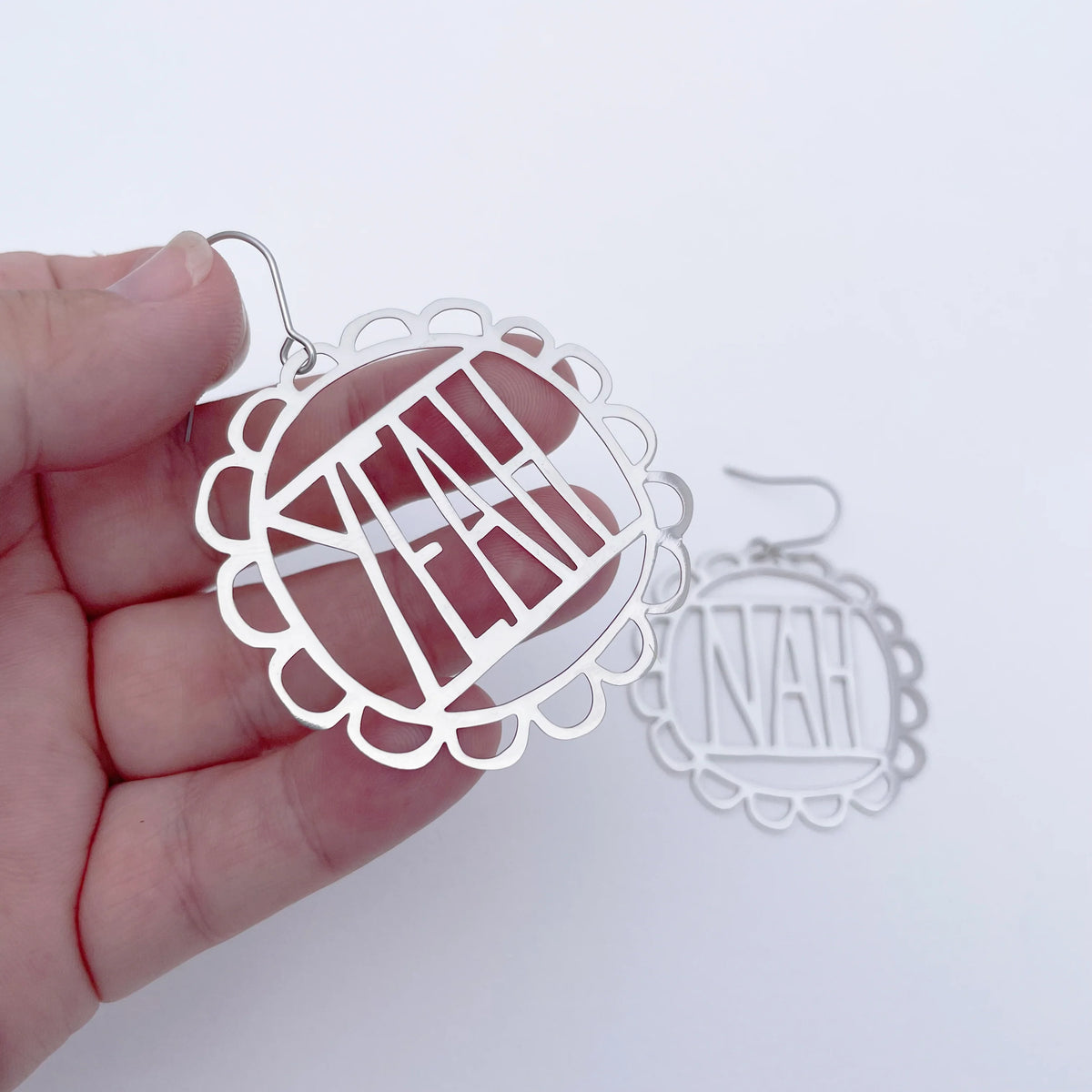 A hand holds a DENZ Yeah Nah earrings in silver with a scalloped border rests on a white surface in the background.