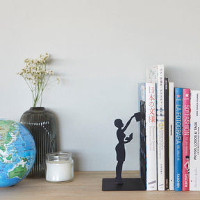 A Balvi black metal bookend styled on a wooden shelf next to books, a globe, a vase of dried flowers, and a candle.