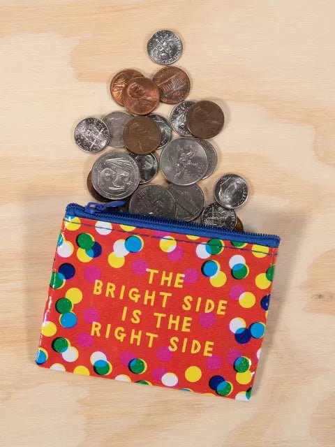 A red Blue Q coin purse covered in colorful polka dots with yellow text reading "THE BRIGHT SIDE IS THE RIGHT SIDE", styled flat-lay with loose coins.