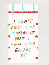 A white folded Blue Q tea towel with red and aqua grid lines, featuring colorful block letters reading "I DON'T FEEL LIKE MAKING IT BUT I FEEL LIKE EATING IT".